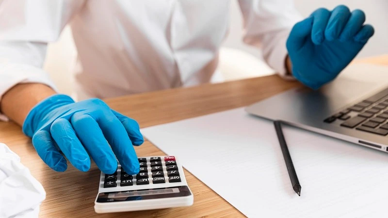 Person an Tisch, von der nur der Oberkörper mit Armen zu sehen ist. Handschuhe an und Taschenrechner, sowie Laptop auf den Tisch liegend, inklusive Papier und Kugelschreiber. Person rechnet etwas am Taschenrechner aus.