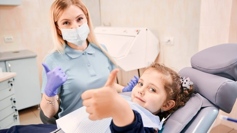 ZFA mit Maske und Kind im Behandlungsstuhl zeigen Daumen hoch – eLearning neue Kinder-Richtlinie Z1-Z6 Zahnarztpraxis | Spitta Akademie